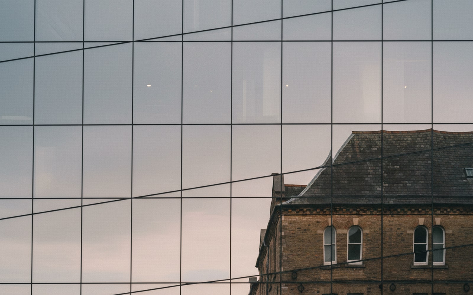 Modern glass office facade reflecting an overcast sky and an older masonry building